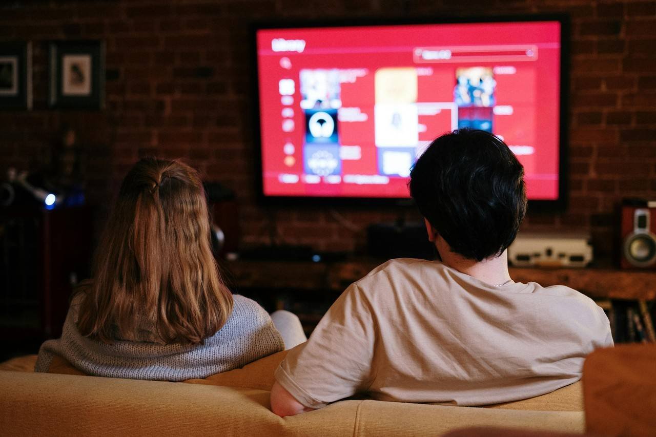 homem e mulher sentados de costas para a câmera assistindo à televisão com som do home theater em uma sala de estar com parede de tijolos