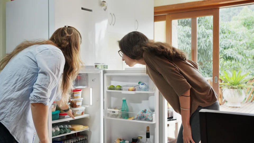 A imagem mostra duas mulheres olhando o interior de um refrigerador aberto em uma cozinha bem iluminada.