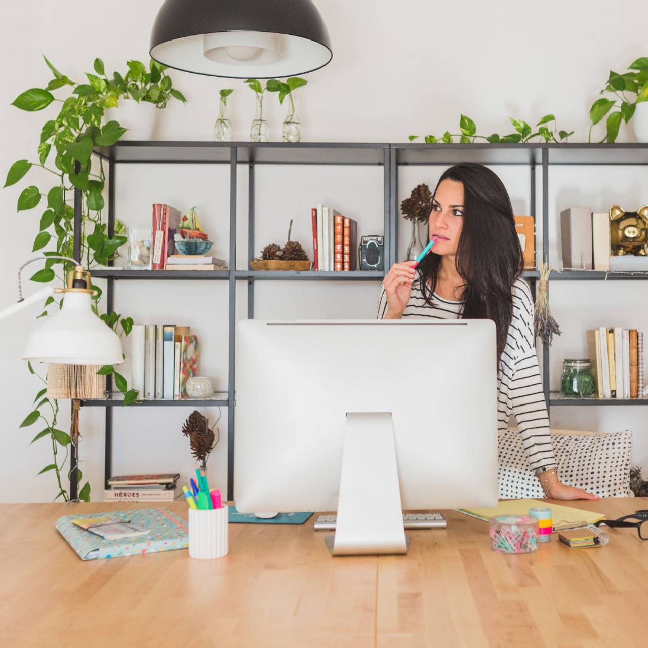 Mulher em um lugar com mesa de madeira com um computador branco e prateleira com livros e plantas