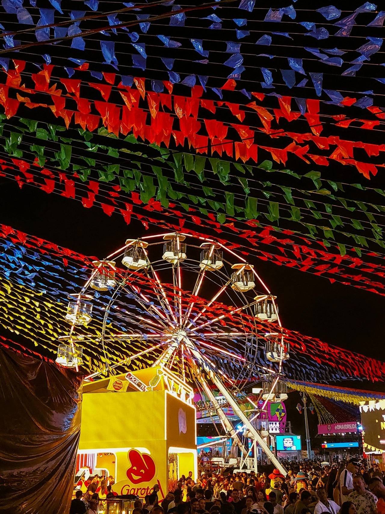 Festa Junina à noite com bandeiras coloridas, barraquinhas de comida, roda gigante e muitas pessoas