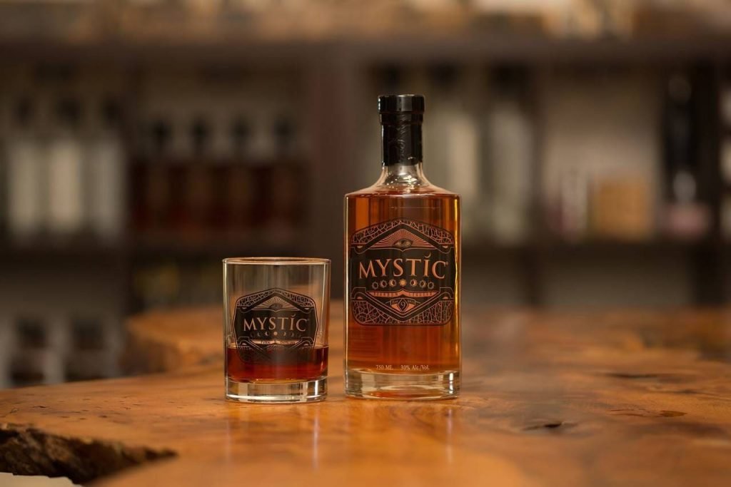 A imagem mostra uma garrafa de licor Mystic com um copo parcialmente preenchido com a bebida.