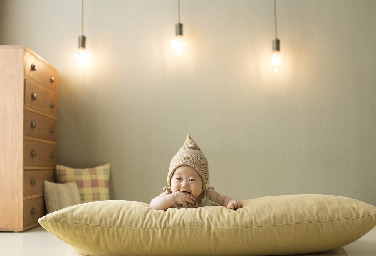 bebê sorrindo com braços apoiados em almofada bege com quarto de bebê de fundo com luminarias pêndulas, almofadas sortidas e cômoda de madeira