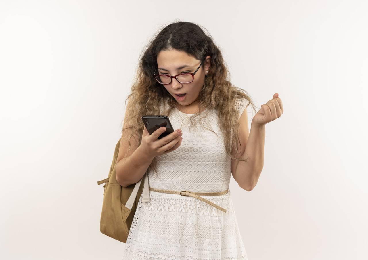 jovem bonita impressionada de colegial usando oculos e bolsa nas costas segurando e olhando para o celular cerrando o punho isolado no branco